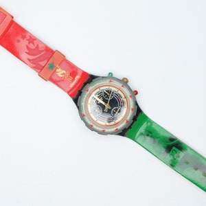 Puede incluir: Un reloj de pulsera rojo y verde con una esfera negra y una esfera blanca. El reloj tiene una función de cronógrafo y presenta una correa roja y verde con un diseño de una antorcha y estrellas. La esfera del reloj tiene el texto "Atlanta 1996" impreso en ella.