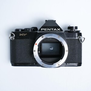 Op de afbeelding: Zwarte Pentax MV camera met zilveren lensvatting, tegen een witte achtergrond. De camerabody heeft een getextureerde grip. Het woord "PENTAX" staat erop.