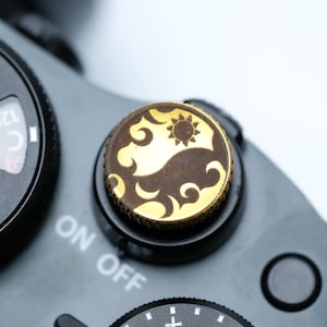 May include: Close-up of a black camera with a gold and brown decorative dial. The dial features a sun and cloud design. The camera's settings dial is visible, with the text "ON OFF" on the camera body.