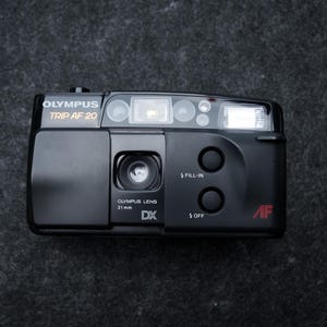 May include: Black Olympus Trip AF 20 camera with a 31mm lens. The camera has a built-in flash and two buttons labeled "FILL-IN" and "OFF". The text "OLYMPUS TRIP AF 20" is printed on the top.