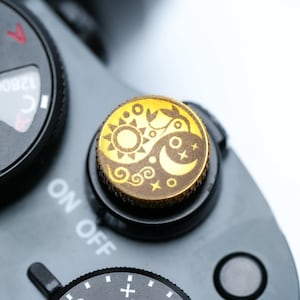 Pode incluir: Uma câmera em close-up com um mostrador dourado com um design de sol e lua. O mostrador está sobre uma base preta com as palavras "ON OFF" visíveis. O mostrador de configurações da câmera também é visível.