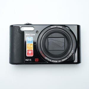 Puede incluir: Cámara digital Ricoh HZ15 negra con una lente prominente y el texto "RICOH LENS". Incluye 16 megapíxeles, zoom óptico 15x y reducción de vibraciones. La cámara está sobre un fondo blanco.