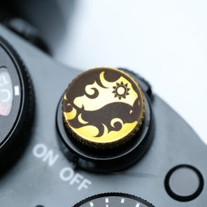 Puede incluir: Primer plano de un dial de cámara con un botón decorativo. El botón presenta un diseño Yin y Yang dorado y marrón con un motivo de sol y nube. El cuerpo de la cámara es negro y el dial está configurado en "ON OFF".