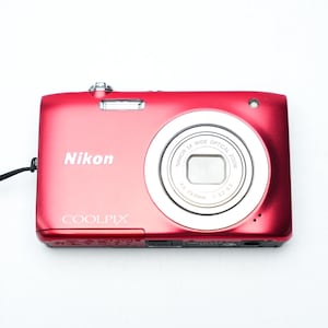 May include: A red Nikon Coolpix digital camera with a silver lens and flash. The camera has the brand name printed on the front and a wrist strap attached. The camera is on a white background.