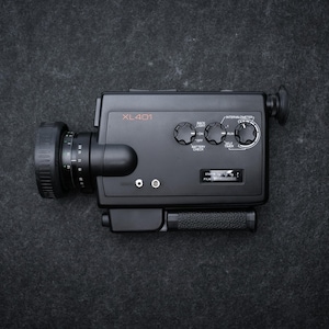 Könnte beinhalten: Schwarze Vintage-XL401-Filmkamera mit Objektiv, Drehknöpfen und Griff. Auf der Kamera steht "XL401". Die Kamera liegt auf einer dunkelgrauen, strukturierten Oberfläche.