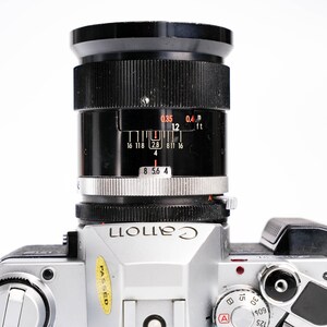 May include: A silver Canon camera with a black lens. The lens has distance markings in feet and meters. The camera body has dials and buttons, and the word "Canon" is printed on the front.