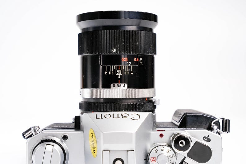 May include: A silver Canon camera with a black lens. The lens has distance markings in feet and meters. The camera body has dials and buttons, and the word "Canon" is printed on the front.