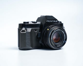 Peut inclure: Appareil photo Pentax noir avec un objectif argenté et le mot "PENTAX" en blanc. L'objectif porte l'inscription "SMC PENTAX-M 1:1.7 50mm JAPAN". L'appareil photo est équipé d'un support de sangle argenté.