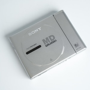 Könnte beinhalten: Ein silberner Sony MD Walkman, Modell MZ-E25, auf einer weißen Oberfläche. Das Gerät trägt das Logo "MD WALKMAN" und den Text "Digital MEGA BASS". Das Gerät ist rechteckig mit abgerundeten Kanten.