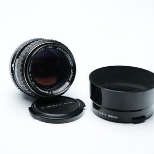 Op de afbeelding: Een zwart en zilveren cameralens met een lensdop en een zwarte zonnekap. De lens is gemarkeerd met "SMC PENTAX-M 1:1.8 50mm" en "ASAHI OPT. CO. JAPAN". De zonnekap is gemarkeerd met "1:2.8-4 100mm".