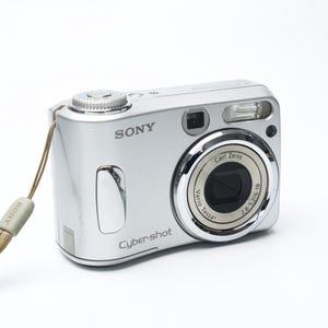 Op de afbeelding: Een zilverkleurige Sony Cyber-shot digitale camera met een Carl Zeiss Vario-Tessar lens met een diafragmabereik van 2,8-5,2/6-18. Het compacte apparaat bevat een ingebouwde flitser, een ontspanknop aan de bovenzijde en een beige polsbandje.