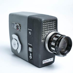 以下が含まれることがあります： グレーとブラックのヴィンテージエルモ8-S 100mmオートアイフィルムカメラ。ブラックのズームレンズ付き。カメラの側面にはシルバーのノブ、上面にはシルバーのダイヤルがあります。