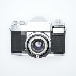 Puede incluir: Una cámara de película de 35 mm plateada y negra con una lente etiquetada como "CONTAFLEX" y "PROTAR 50 mm f/2.8".