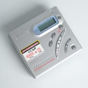 Könnte beinhalten: Silberner Sony MiniDisc Walkman-Player mit kleinem LCD-Bildschirm und mehreren Bedientasten. Das Gerät hat einen Aufkleber mit der Aufschrift "MDLP" und "4 CDs". Der Text "Recording MD Walkman" ist ebenfalls sichtbar.