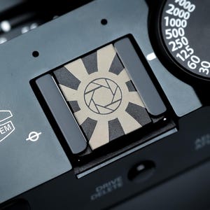 Op de afbeelding: Close-up van een zwarte camerabody met een zilveren lenssysteemlabel. De camera heeft een draaiknop met witte cijfers en een flitsschoen met een sunburst-ontwerp. De tekst "FUJINON LENS SYSTEM" is zichtbaar.