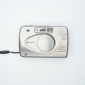 Op de afbeelding: Een zilveren Minolta Vectis 300 filmcamera met een zwarte band. De camera heeft een ingebouwde flitser en een datumtempel.