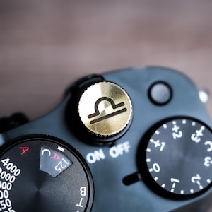 May include: Close-up of a black camera with gold accents. The camera features a gold dial with the Libra zodiac symbol, and various control dials with white markings. The text "ON OFF" is visible.