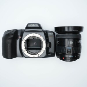 May include: A black Minolta Dynax 300si Super SLR camera with a black Minolta AF 28-80mm lens attached. The camera has a silver ring around the lens mount.
