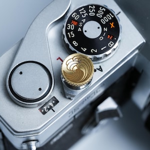 May include: Close-up of a silver camera body with black and gold dials. The top features a black dial with white numbers and a gold-colored button with a wave design. The camera has a vintage aesthetic.