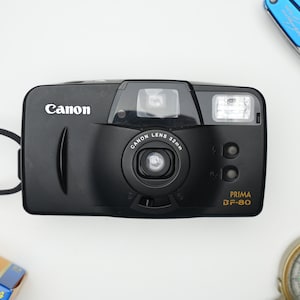 May include: A black Canon Prima BF-80 35mm film camera with a black strap. The camera has a lens with a 32mm focal length.