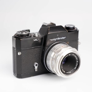 May include: A black Voigtländer VSL 1 35mm film camera with a silver lens. The camera has a black leatherette covering and a silver metal lens.