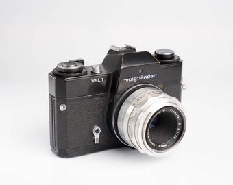 Peut inclure: Un appareil photo argentique noir Voigtländer VSL 1 35 mm avec un objectif argenté. L'appareil photo est recouvert de simili cuir noir et possède un objectif en métal argenté.