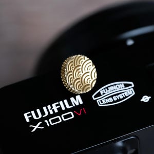 Op de afbeelding: Close-up van een zwarte Fujifilm X100VI camera met een goudkleurig, cirkelvormig accessoire. Het accessoire heeft een herhalend golfpatroon. De camera heeft witte tekst met "FUJIFILM X100VI" en "FUJINON LENS SYSTEM".