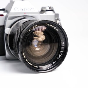 May include: A silver Canon AL-1 camera with a black Tamron 28mm lens. The lens has the text "AUTO TAMRON 1:2.8 28mm" and "LENS MADE IN JAPAN" printed on it. The camera is on a white background.