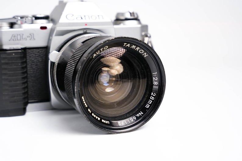May include: A silver Canon AL-1 camera with a black Tamron 28mm lens. The lens has the text "AUTO TAMRON 1:2.8 28mm" and "LENS MADE IN JAPAN" printed on it. The camera is on a white background.