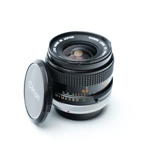 Puede incluir: Un objetivo Canon FD 35mm f/2.8 con cuerpo negro y anillo plateado. La tapa del objetivo está quitada y apoyada junto al objetivo. La distancia de enfoque es de 0,3 metros.