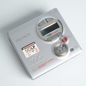 Op de afbeelding: Zilveren Sony MD Walkman MZ-R410. De draagbare audiospeler heeft een digitaal display, bedieningsknoppen en de tekst "MDLP TYPE-R". Het apparaat heeft een sticker met de tekst "MDLP" en "400%".