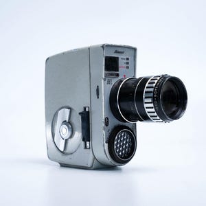 May include: A silver and black vintage Bauer 8SL Super 8 film camera with a black lens and a silver rewind knob. The camera has a built-in light meter and a viewfinder.
