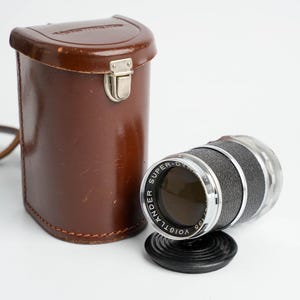 Peut inclure: Un objectif d'appareil photo vintage avec un étui en cuir marron. L'objectif est noir et argenté avec le texte "SUPER-DIAPHRAGM" et "VOIGTLANDER". L'étui a un fermoir en métal et une sangle en cuir.
