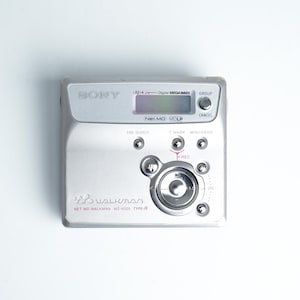 Könnte beinhalten: Silberner Sony Walkman Net MD MZ-N505 Type-R Digital-Musikplayer. Er verfügt über einen kleinen LCD-Bildschirm, Steuertasten und einen Lautstärkeregler. Das Gerät hat das Walkman-Logo und andere Texte auf der Vorderseite.
