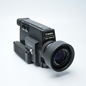 May include: A black Canon Auto Zoom 512 movie camera featuring a prominent lens with a circular hood, a textured grip, and top-mounted control buttons. The device displays the brand name Canon and the model text Auto Zoom 512 on the side.