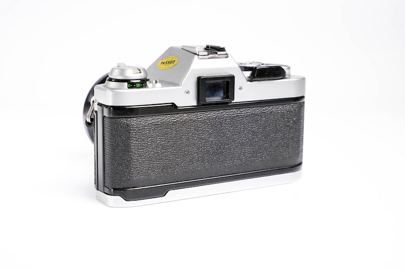 May include: Silver and black vintage camera with a textured grip. The camera has a lens, viewfinder, and a yellow "PASSED" sticker. The camera strap is black. The camera is on a white background.
