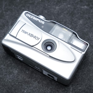 May include: Silver Olympus Trip XB401 camera. The camera has a rectangular shape with rounded edges. The text "OLYMPUS" is printed on the top, and "TRIP XB401" is printed on the front. The camera has a lens and a flash.