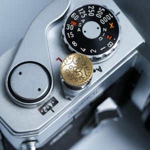 Può includere: Primo piano di una fotocamera vintage argentata con quadranti neri e un pulsante color oro. I quadranti mostrano numeri e impostazioni per l'apertura e la velocità dell'otturatore. Il design della fotocamera è classico e dettagliato.