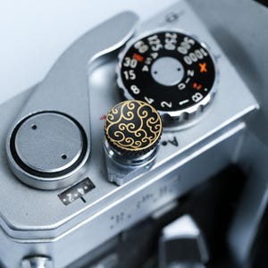 Può includere: Primo piano di una fotocamera vintage argentata con quadranti neri e una manopola decorativa nera e oro. I quadranti della fotocamera presentano segni bianchi e rossi. La manopola decorativa ha un motivo a spirale dorato.