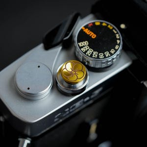 Puede incluir: Una cámara plateada con una correa de cuero negra y un botón dorado con un diseño floral. La cámara tiene un dial negro con números blancos y la palabra "AUTO" impresa en naranja.