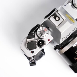 May include: Silver Canon camera with a black lens, featuring a dial with speed settings from 15 to 1000. The lens has measurement markings. The camera is set against a white background.