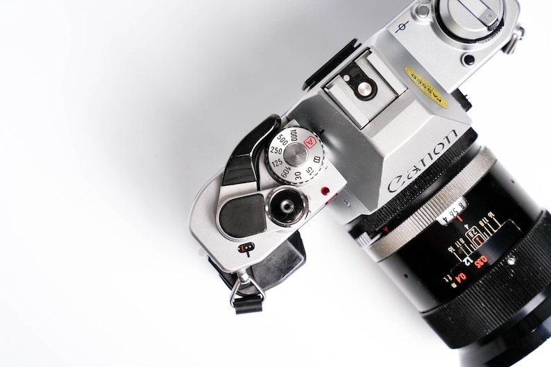 May include: Silver Canon camera with a black lens, featuring a dial with speed settings from 15 to 1000. The lens has measurement markings. The camera is set against a white background.