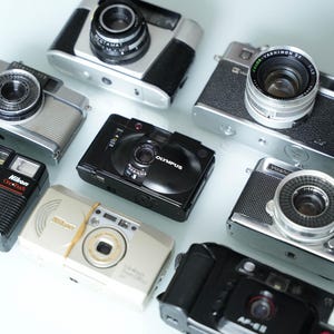 Puede incluir: Una colección de cámaras de película vintage y modernas, incluyendo una Olympus plateada, una Nikon negra, una Yashica plateada y una Canon negra.
