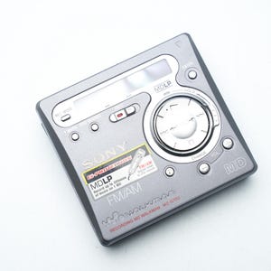 Könnte beinhalten: Ein silberner tragbarer digitaler Musikplayer Sony MD Walkman MZ-G750. Das Gerät hat einen kreisförmigen Drehregler, einen kleinen LCD-Bildschirm und mehrere Tasten. Das Gerät hat einen Aufkleber mit der Aufschrift "MDLP FM/AM".