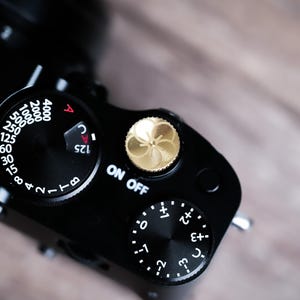 Puede incluir: Primer plano de una cámara negra con varios diales y ajustes. La cámara presenta un botón dorado y texto blanco que indica ajustes como ISO, velocidad de obturación y encendido. El fondo es una superficie de madera borrosa.