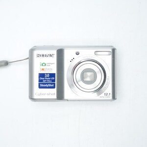 May include: A silver Sony Cyber-shot digital camera with a wrist strap. The front features a lens, a flash, and labels including 12.1 Mega Pixels, 3.0 Clear Photo LCD 3.0/7.5cm, SteadyShot, Intelligent Auto, and Smile Shutter technology.