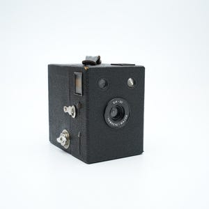 May include: A black vintage box camera with a silver lens and a silver key on the side. The camera has the text "Six-20" and "Harfette Major" printed on it.
