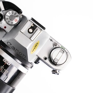 May include: A silver Canon camera with a black lens, viewed from above. The camera has various dials and settings visible, including a speed dial and a focus ring. A yellow "PASSED" sticker is on the body.
