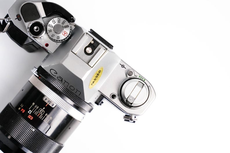 May include: A silver Canon camera with a black lens, viewed from above. The camera has various dials and settings visible, including a speed dial and a focus ring. A yellow "PASSED" sticker is on the body.