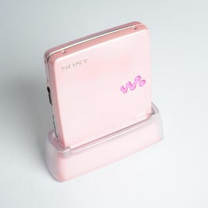 Könnte beinhalten: Ein rosa Sony MiniDisc-Player mit einer passenden rosa Halterung. Der Player hat das Sony-Logo und ein stilisiertes 'W'-Logo in dunklerem Rosa. Das Gerät steht vor einem weißen Hintergrund.